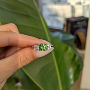 Può includere: Un anello color argento con una pietra centrale rotonda di un verde vibrante, affiancata da due pietre trasparenti rettangolari più piccole. La montatura è metallica con finitura lucida e presenta un design a tre pietre con griffe.