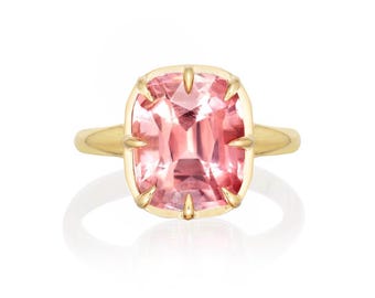 May include: A gold ring featuring a large, cushion-cut pink gemstone. The gemstone is set in a gold bezel with claw prongs. The ring has a simple, polished gold band. The ring is displayed on a white background.
