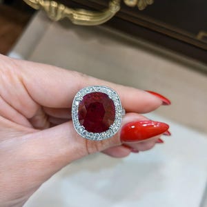 Puede incluir: Un anillo con un gran rubí de corte rectangular, rodeado por un halo de pequeños diamantes claros. El rubí es de color rojo intenso, y la montura del anillo parece ser de plata u oro blanco. Una pieza llamativa.