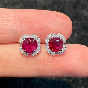 May include: A pair of stud earrings featuring a central cushion-cut deep red ruby surrounded by a halo of small round and baguette-cut white diamonds set in a silver-toned metal frame, creating a square-shaped design with rounded corners.