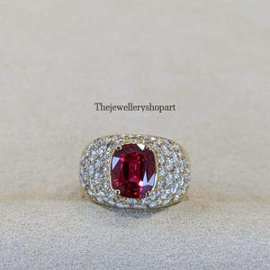 Può includere: Un anello d'oro con una grande gemma rosso scuro a taglio cuscino al centro, circondata da un alone di gemme chiare più piccole. Anche la fascia dell'anello è adornata da gemme più piccole. Il testo "Thejewelleryshopart" è visibile.