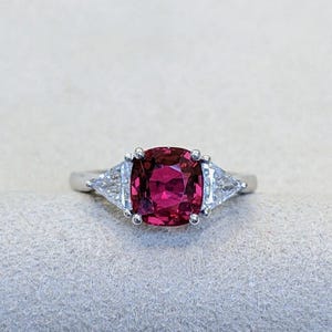 Op de afbeelding: Een zilveren ring met een grote, kussenvormige, dieprode edelsteen als middelpunt. Twee kleinere, driehoekige, heldere edelstenen flankeren de hoofdsteen. De ring is geplaatst tegen een neutrale achtergrond, waardoor de levendige kleuren van de edelstenen worden benadrukt.