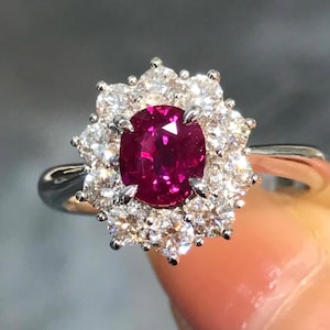 Può includere: Un anello d'argento con un grande rubino ovale al centro, circondato da un alone di diamanti rotondi più piccoli. Il rubino è di un rosso vibrante e i diamanti brillano intensamente. L'anello è tenuto da una mano.