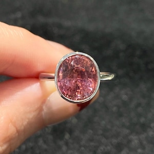 Puede incluir: Un anillo de plata con una gran gema rosa de talla ovalada. La gema está engastada en un bisel y el anillo tiene una banda simple y pulida. La piedra rosa tiene un aspecto ligeramente translúcido. Joyería.