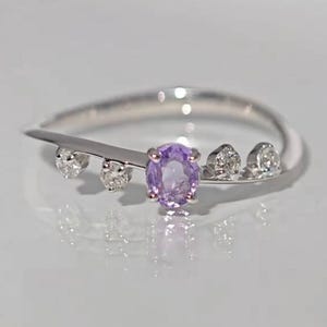 May include: A silver ring featuring a central oval-cut purple gemstone, flanked by three smaller, round, clear gemstones. The ring has a delicate, flowing design, with the gemstones set on a slender band. The ring is displayed on a white surface.