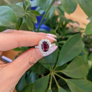 May include: An oval-cut, deep red gemstone ring with a halo of smaller, clear stones. The ring is set in a gold band. The background features green foliage. The text "Thejewelleryshopart" is visible.
