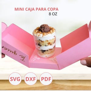 Puede incluir: Una caja de regalo rosa con el texto "Mini Caja Para Copa" y "8 OZ" en blanco. En el interior, se ve un postre en capas en una taza transparente. La caja está abierta, revelando el postre y las palabras "Te quiero" en dorado.