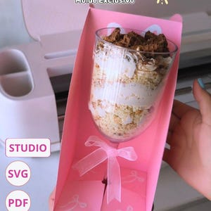Puede incluir: Una caja de regalo rosa contiene una copa de postre con ingredientes en capas: una base clara, crema blanca y trozos de chocolate negro. La caja está atada con una cinta y tiene el texto "CAJA DE COPA" y "Molde Exclusivo".