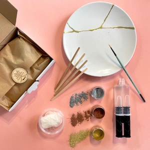Op de afbeelding: Een DIY-kit voor het maken van een gebarsten gouden bord. De kit bevat een wit keramiek bord met gouden barsten, epoxyhars, een klein penseel, houten stokjes en verschillende gekleurde poeders en glitter.