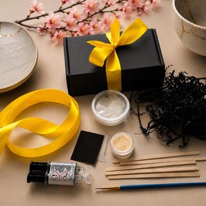 Op de afbeelding: Een Kintsugi-reparatieset met een zwarte doos vastgebonden met een gele lint, een grijze kom en een witte kom met gouden accenten. De set bevat lijm, poeder, een penseel en houten stokjes. Roze bloemen en geel lint zijn ook aanwezig.