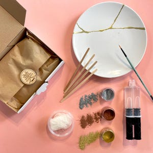 Op de afbeelding: Een Kintsugi-reparatieset met een witte keramische plaat met goudkleurige reparatielijnen. De set bevat een doos, poeders, een penseel, roerstaafjes en een lijmdispenser. De items zijn gerangschikt op een roze oppervlak.