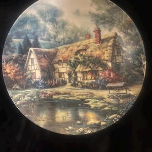 May include: A round ceramic plate with a painting of a thatched-roof cottage with a pond in front of it. The cottage is surrounded by trees and flowers. The painting is in a soft, muted color palette.