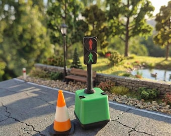 Può includere: Semaforo in miniatura verde e nero con segnale di attraversamento pedonale, accanto a un cono arancione e bianco. La scena è ambientata su una strada asfaltata in miniatura con un parco sullo sfondo.