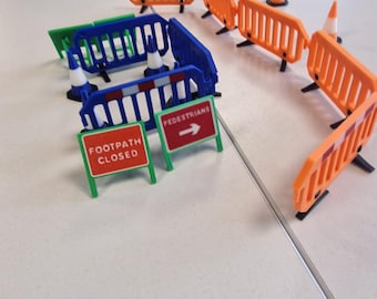 May include: Miniature construction site setup with orange and blue barricades, traffic cones, and signs. Signs read "Footpath Closed", "Pedestrians", and "Works Exit". The scene is set on a light-colored surface.
