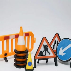 Può includere: Una collezione di segnali stradali e attrezzature di sicurezza per lavori stradali in miniatura. Include coni arancioni e bianchi, una barriera giallo-arancio e vari segnali stradali con grafica in bianco e nero.