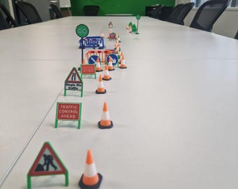 Può includere: Una scena in miniatura di lavori stradali con coni arancioni e bianchi, segnali stradali e una barriera blu. I segnali mostrano testo come "Traffico a fila unica" e "Controllo del traffico in arrivo". La scena è ambientata su un tavolo bianco.
