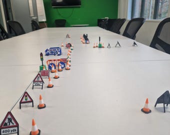 Può includere: Scena in miniatura di lavori stradali con coni arancioni e bianchi, segnali stradali e un semaforo. I segnali includono "Single File Traffic" e "400 yds". La scena è ambientata su una superficie bianca, con un muro verde e una finestra sullo sfondo.