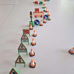 Op de afbeelding: Miniatuur verkeersborden en oranje en witte verkeerskegels op een witte ondergrond. De borden waarschuwen voor wegwerkzaamheden, verkeerslichten en afstanden. De kegels zijn oranje en wit. De borden tonen 'Werktoegang' en 'Enkel bestandsverkeer'.