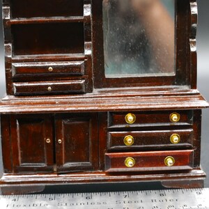 May include: Miniature wooden dresser with a mirror, shelves, and drawers. The dresser is stained a dark brown color with gold-colored knobs. The mirror is rectangular and has a slightly cloudy appearance. The dresser is a small scale, likely for a dollhouse.