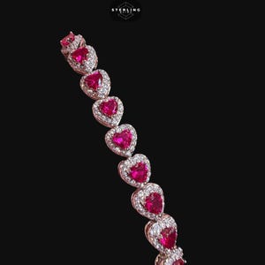 May include: A bracelet featuring a series of heart-shaped red stones, each surrounded by a halo of small clear stones set in a metallic band. The logo STERLING BLING is visible at the top. The design consists of repeating heart links in a row.