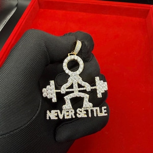 May include: A gold-colored pendant featuring a weightlifter design encrusted with clear gemstones. The pendant includes the phrase "NEVER SETTLE" in a matching gemstone font. The pendant is held against a red background.