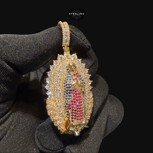 May include: Gold-toned pendant featuring a religious figure, adorned with colorful gemstones. The pendant has a halo design with a central figure in red, blue, and gold. The pendant is held by a gloved hand against a dark background.