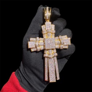 May include: A gold-colored cross pendant encrusted with numerous small, sparkling gemstones. The cross has a detailed, layered design with a bail at the top for a chain. The pendant is held against a black background.