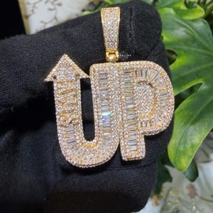 May include: Gold-toned pendant spelling out "UP" with an arrow pointing upwards. The letters are encrusted with small, sparkling stones. The pendant is held against a black background with green foliage visible.