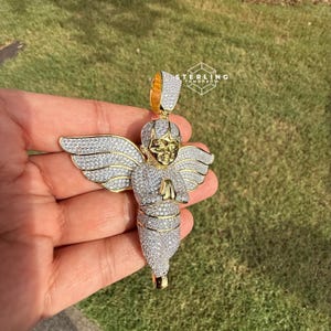 May include: A gold-toned angel pendant with a halo and wings, encrusted with sparkling white gemstones. The angel is depicted with folded hands in prayer. The pendant is held against a backdrop of green grass.