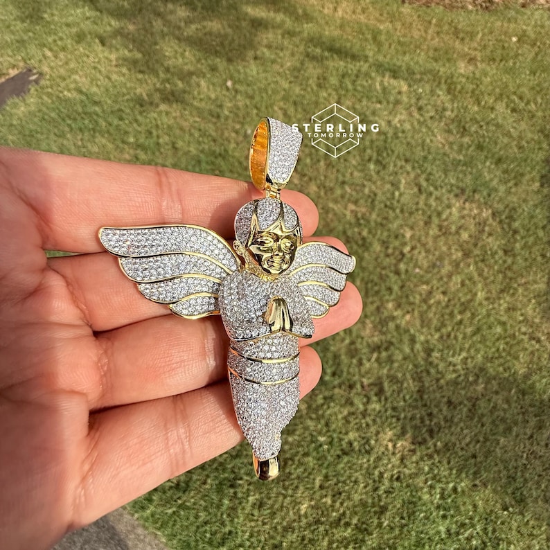 May include: A gold-toned angel pendant with a halo and wings, encrusted with sparkling white gemstones. The angel is depicted with folded hands in prayer. The pendant is held against a backdrop of green grass.
