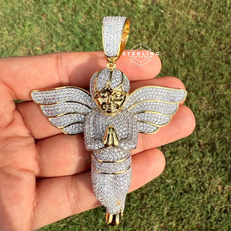 May include: Gold-toned angel pendant encrusted with sparkling white gemstones. The angel has outstretched wings and clasped hands. The pendant's bail is also gold-toned and covered in gemstones. The angel's face is a contrasting gold color.