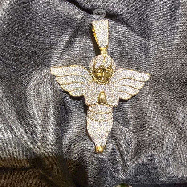 May include: A gold-toned angel pendant encrusted with sparkling white gemstones. The angel has outstretched wings and clasped hands in a praying gesture. The pendant is suspended from a matching bail.