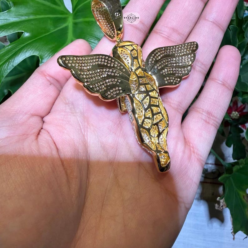 May include: A gold-colored angel pendant with detailed wings and a textured body. The pendant is adorned with small, sparkling accents, and is suspended from a matching gold bail. The design is intricate and eye-catching, suitable for jewelry.