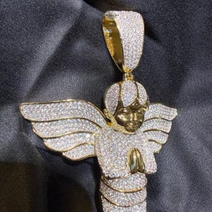May include: A gold-toned angel pendant with sparkling white gemstones. The angel has outstretched wings and clasped hands. The pendant is suspended from a gold bail, and the overall design is intricate and detailed.