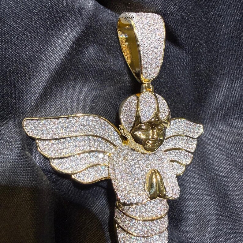 May include: A gold-toned angel pendant with sparkling white gemstones. The angel has outstretched wings and clasped hands. The pendant is suspended from a gold bail, and the overall design is intricate and detailed.