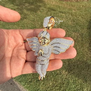 May include: A gold-toned angel pendant with a halo and outstretched wings, encrusted with sparkling white gemstones. The angel is depicted with hands clasped in prayer. The pendant is held against a backdrop of green grass.