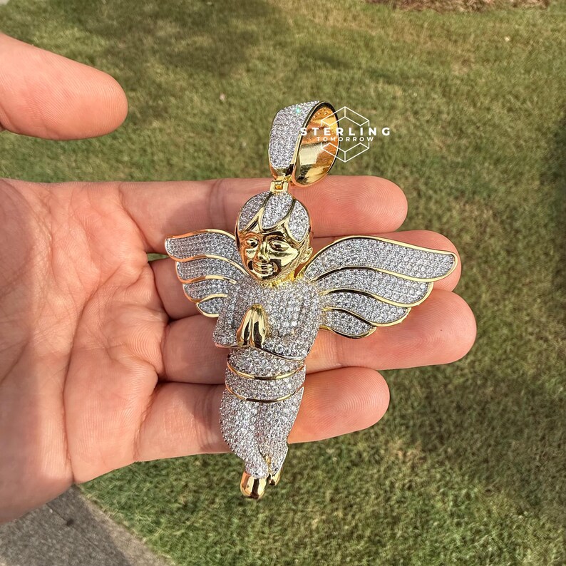 May include: A gold-toned angel pendant with a halo and outstretched wings, encrusted with sparkling white gemstones. The angel is depicted with hands clasped in prayer. The pendant is held against a backdrop of green grass.