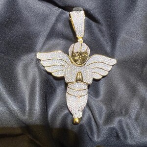 May include: A gold-toned angel pendant encrusted with sparkling white gemstones. The angel has outstretched wings and clasped hands in a prayer position. The pendant is suspended from a matching bail.