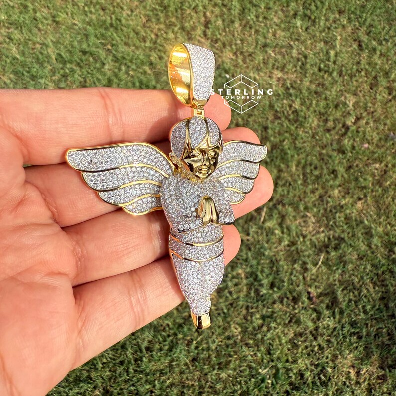May include: Gold-toned angel pendant encrusted with sparkling white stones. The angel has outstretched wings and clasped hands. The pendant is held by a gold bail, and the design is set against a green background.