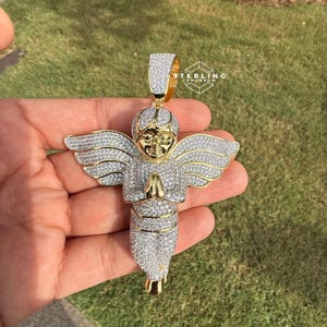 May include: A gold-toned angel pendant encrusted with clear crystals. The angel has outstretched wings and clasped hands. The pendant is held against a background of green grass. The text "STERLING TOMORROW" is visible.