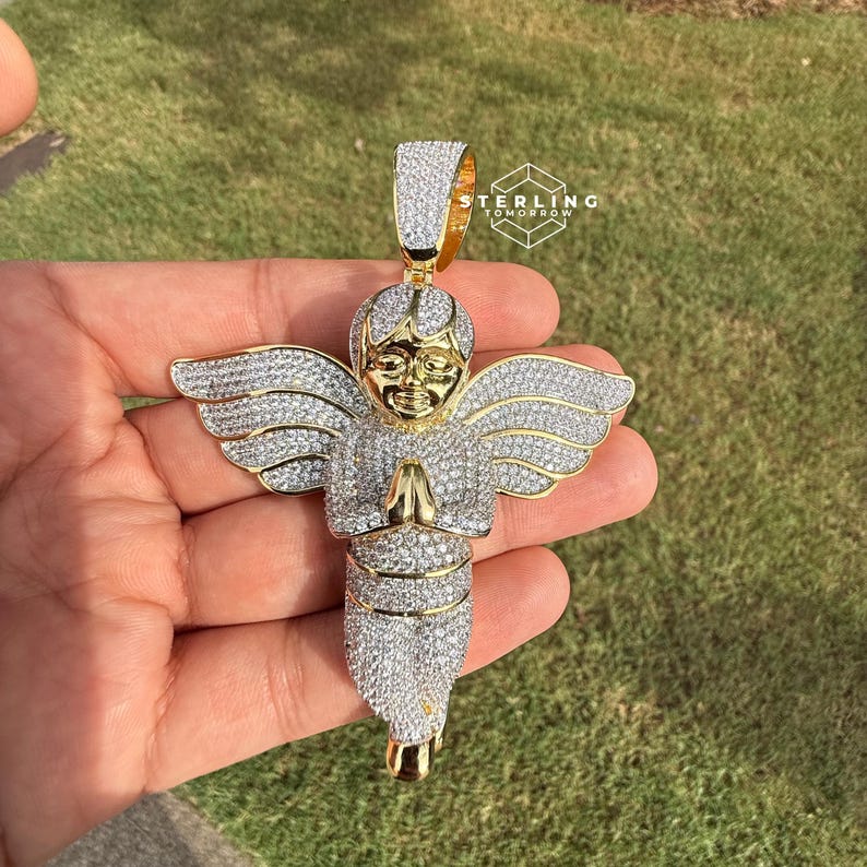May include: A gold-toned angel pendant encrusted with clear crystals. The angel has outstretched wings and clasped hands. The pendant is held against a background of green grass. The text "STERLING TOMORROW" is visible.