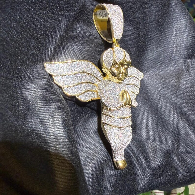 May include: Gold-toned angel pendant with a halo and outstretched wings, encrusted with sparkling white gemstones. The angel is depicted with hands clasped in prayer. The pendant hangs from a gold bail.