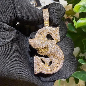May include: A gold-toned pendant in the shape of a dollar sign, encrusted with sparkling white and baguette-cut gemstones. The pendant has a stylized shark-like design with a smiling mouth. The bail is also covered in gemstones.