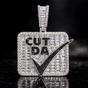 May include: A square pendant featuring a silver-tone metal frame encrusted with clear baguette and round stones. The center displays the black text CUT DA with a large black checkmark symbol, all set against a background of clear stones.