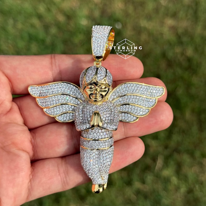 May include: A gold-toned angel pendant with sparkling white gemstones. The angel has outstretched wings and clasped hands. The pendant is held against a green background.