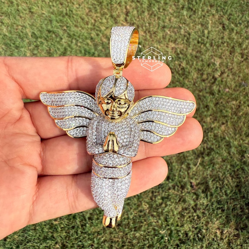 May include: A gold-toned angel pendant encrusted with sparkling white stones. The angel has outstretched wings and clasped hands, with a gold face and a gold bail. The pendant is held against a backdrop of green grass.