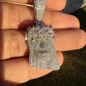 May include: A silver pendant featuring a detailed depiction of Jesus' face, encrusted with numerous small, sparkling stones. The pendant has a bail for a chain and is held against a blurred background.