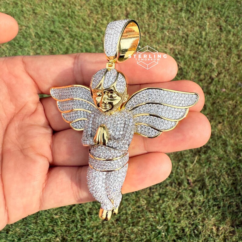 May include: A gold-toned angel pendant encrusted with clear crystals. The angel is depicted with folded hands and outstretched wings. The pendant has a bail for a chain and is set against a green background.