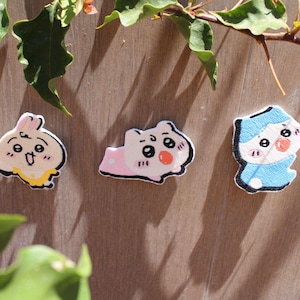 Puede incluir: Tres parches bordados con diseños de personajes de dibujos animados. Los parches presentan un conejito, un hámster y un personaje con un atuendo azul. Cada parche tiene un contorno negro y está sobre un fondo de madera.