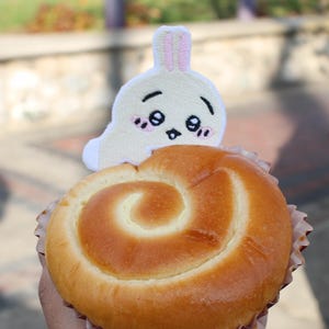 Puede incluir: Un pastel dorado en forma de espiral, en una taza de papel, coronado con un pequeño conejito de peluche blanco con una cara linda. El pastel se sostiene en una mano, con un fondo borroso al aire libre.